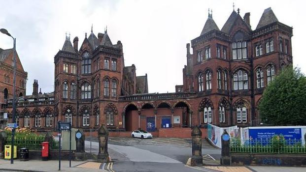 Leeds General Infirmary is pictured. It is a Gothic-style red brick building with several towers. It features tall, narrow windows and arches. A white car is parked in front of the hospital.
