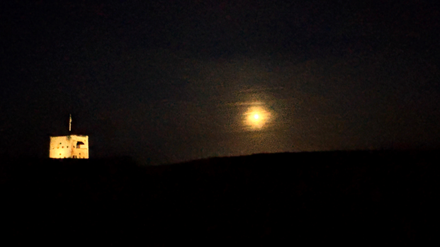 A bright orange moon lighting up a very dark night There is a large building, part of which is lit up. The rest of the image is in complete darkness. 