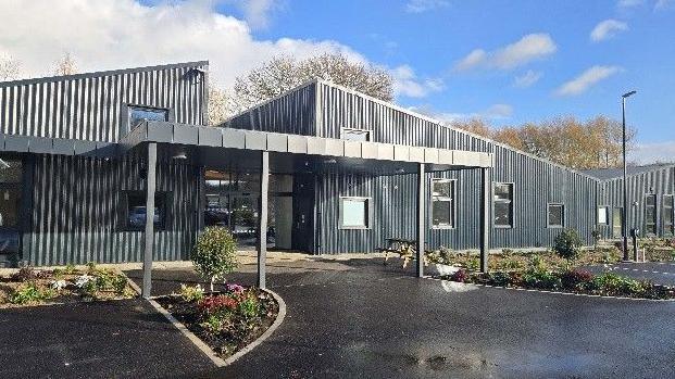 A two storey modern building with a slanted roof and a entrance canopy. It is clad in grey and there are beds with plants in them outside the front of the building. Behind the building are treetops and a blue sky.