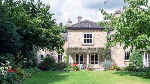 A large stone house with two doors leading to a garden with grass and trees along the side. There are red and white flowers in the beds beside the grass.