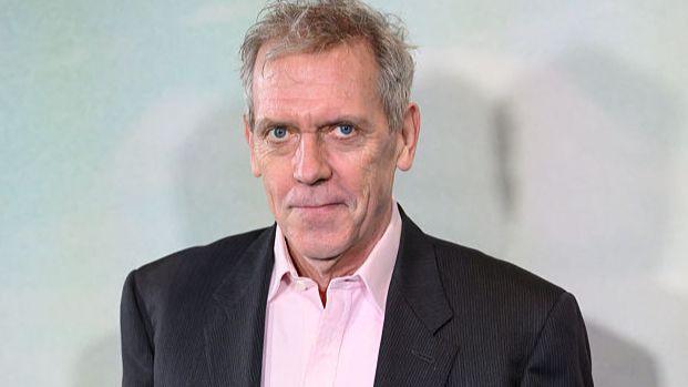 Hugh Laurie smiling at the camera. He is wearing a pink shirt and black blazer