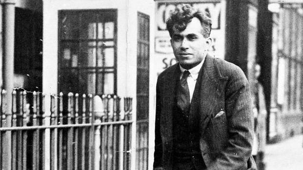 Black-and-white picture of Dr Buck Ruxton, wearing a three-piece suit and tie. He is walking next to railings and there is a row of shops in the background.