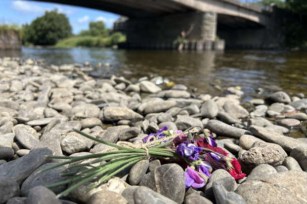 Stones at the side of the River Wye with a bunch of flowers left on them. The flowers are in focus and the river and bridge are blurred.