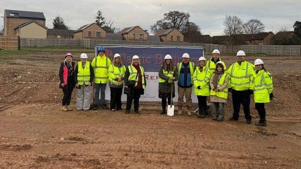 A group of people in high visibility jackets and hard hats on an empty field building site, one holding a spade