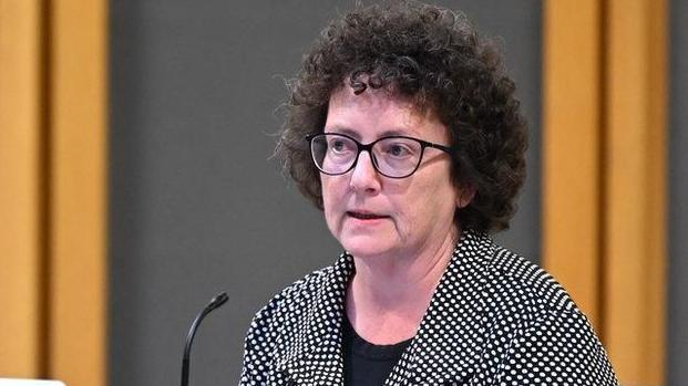 Elin Jones who has dark curly hair, stood at a podium and wearing a black and white polkadot jacket and black top