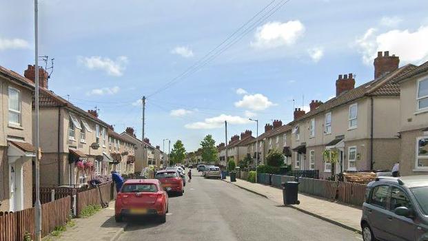 There are rows of terraced houses on either side of Fairbrook Drive in Birkenhead North. There are are number of cars parked on the residential street. 