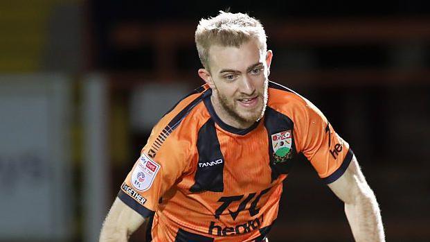 Callum Stead runs after the ball in his amber and black Barnet home shirt. He is looking down at the ball which is out of the picture. 