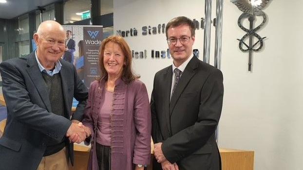 Mark Barnish is wearing a dark suit and grey jumper and he is shaking hands with Grace Jordan who is wearing a purple outfit and has long, brown hair.  Andrew Micklin is wearing a dark grey suit and glasses and is stood with his hands together smiling at the camera.