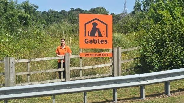 Claire Sparkes is pictured standing in front of an orange Gables sign. She is standing to the left of the sign, behind the fence which is overgrown with plants and trees.