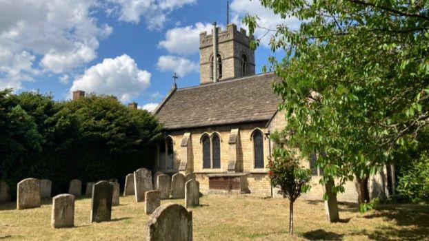 The churchyard with a cemetery in front and the church building in the background, surrounded by trees on either side.