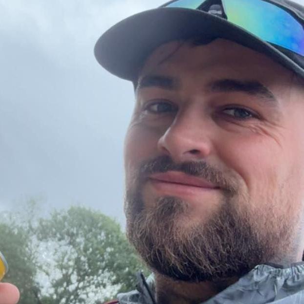 Jordan has blue eyes and a dark brown beard. He is wearing a black cap and blue reflective sunglasses on top of his cap. He is standing outside with trees in the distance. The sky is grey.