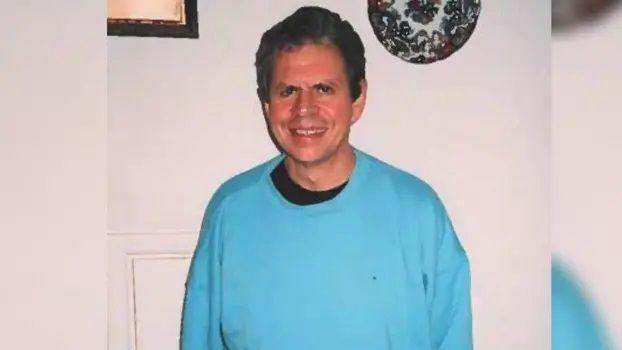 Stephen Ridgeway wearing a black t-shirt underneath a blue jumper. He has short black curly hair and is smiling at the camera. He is standing in front of a white wall which has a decorative plate and a picture frame displayed on it.
