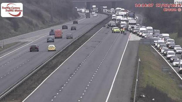 CCTV image showing queuing traffic on a motorway and onto an exit road. A vehicle and two people in hi-vis jackets can be seen in front of blocks placed on the road.
