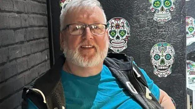 Kevin Price, who had white hair and a beard, is photographed wearing dark glasses. He smiles as he is photographed sitting in a room with black wallpaper, wearing a gilet on top of a blue t-shirt.