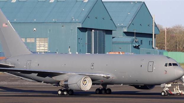 A USAF KC-46A Pegasus tanker aircraft pictured at Prestwick Airport. The grey aircraft rests on tarmac with two aircraft hangers pictured behind it.