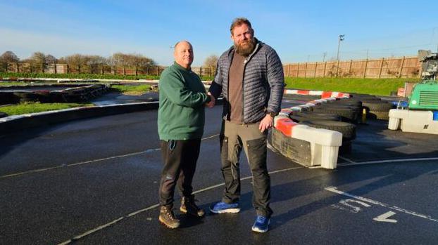 A shorter man with a bald head and green fleece shakes hands with a taller man with a bushy beard and navy jacket. The pair are standing on a karting track on a sunny day.