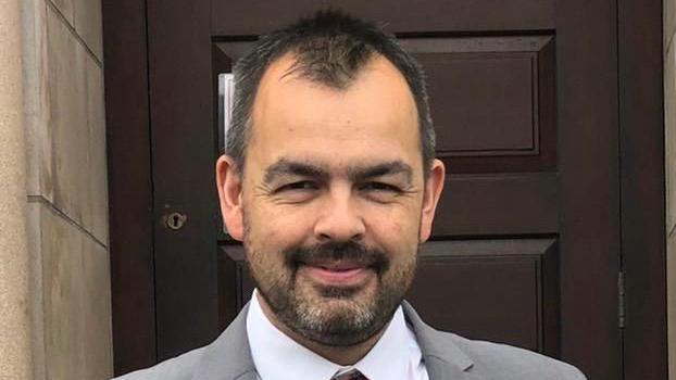 Mr Kane with short black hair and facial hair. He smiles at the camera as he wears a grey suit with a white shirt and red patterned tie.