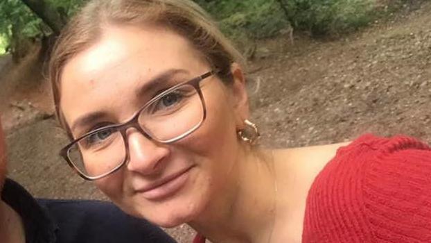 Isobella Knight with blonde hair tied back, wearing brown-framed glasses and a red top. She also has a large gold earring in her left earlobe. There is a road or footway behind her and part of a tree to the left.