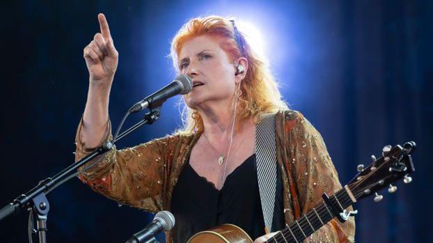 Eddi Reader, who has ginger, shoulder length hair, holding a guitar in her left hand and pointing upwards with her right as she speaks/sings into a microphone with a bright white light behind her 