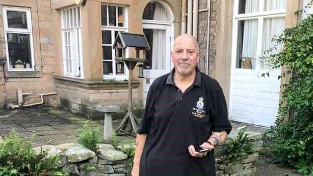 Peter Wright, in a black t-shirt and trousers, is standing in front of Shotley Park and holding a mobile phone. He has grey beard. The frontage of the stone building and its white doors is visible behind him. 