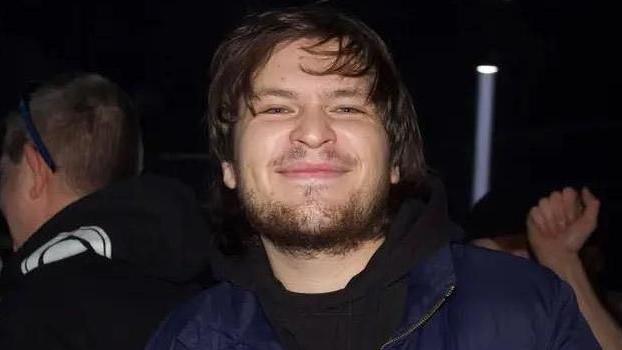 Ezra Tilford-Key in what appears to be a nightclub or party. He is smiling. He has a full beard and moustache, and has medium-length dark hair. He is wearing a black t-shirt and a blue jacket. A light shaft from a spotlight can be seen behind him.
