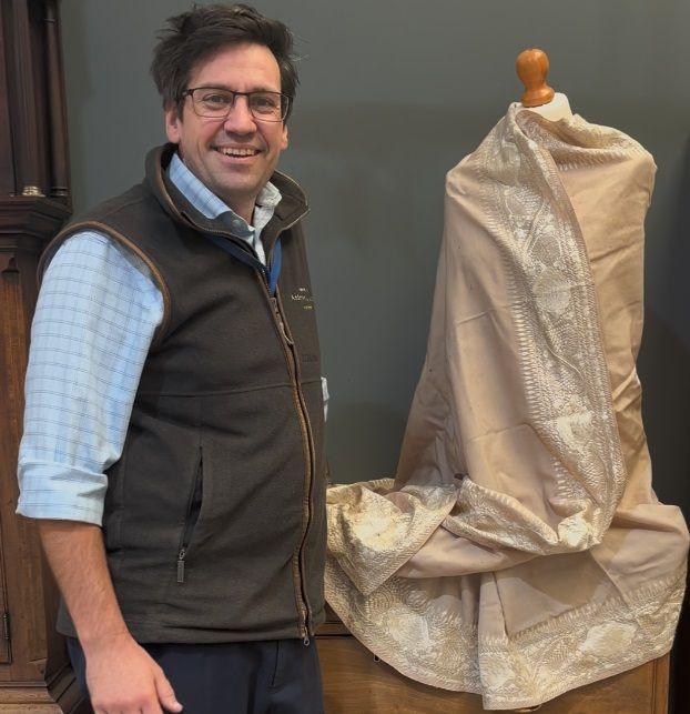 Fred Wyrley-Birch, with dark hair and glasses, wearing a blue checked shirt and brown gilet, standing next to the shawl on a mannequin.