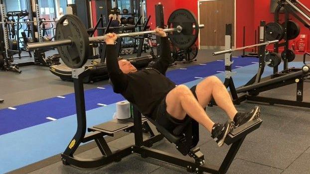 A man dressed in a black t-shirt and black shorts lying on a weight bench lifting weights on a barbell. He is in a weights room surrounded by other equipment.