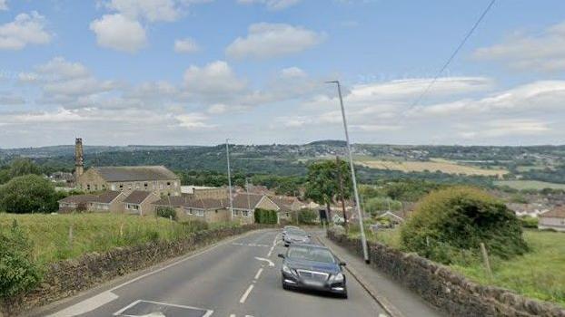 A road with a dry stone wall on either side, surrounded by fields. In the backdrop are houses, buildings and hills. There are three cars shown being driven on the road.