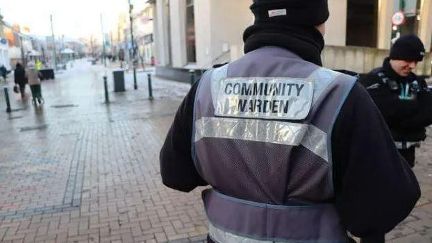 A man stands with his back to the camera. He is wearing a jacket with the wording "community warden" on the back. A high street can be seen in the background. 