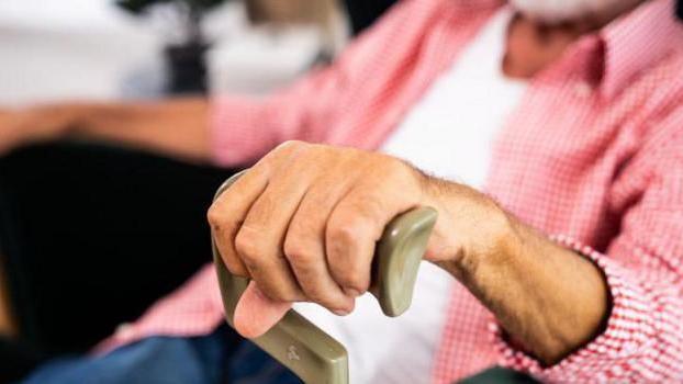 A close-up image of an elderly person's hand gripping the top handle of a walking stick. They appear to be sitting down while wearing a red and white chequered shirt over a white T-shirt.