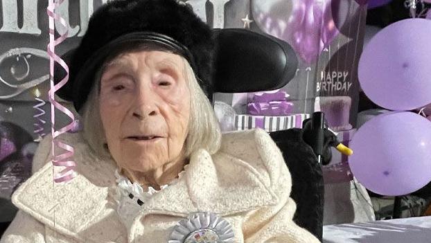 Dorothy Smith marking her 105th birthday at Bottisham Airfield. She has shoulder-length silver hair and is seated, wearing a cream coloured coat with black gloves, holding a grey blanket over her lap. Behind her is a grey sign saying "Happy Birthday" in white letter and to her left are purple balloons