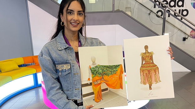 Daya Bhatti holds illustrations of her designs. She has a denim jacket on and has long black hair.