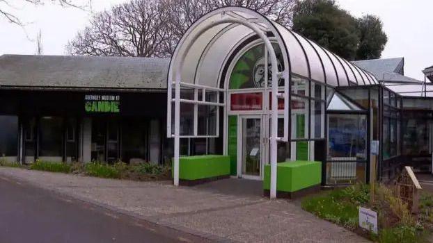 Guernsey Museum entrance which is an oval structure that is largely glass with a green base and white painted frame. The photo is taken on a cloudy day.
