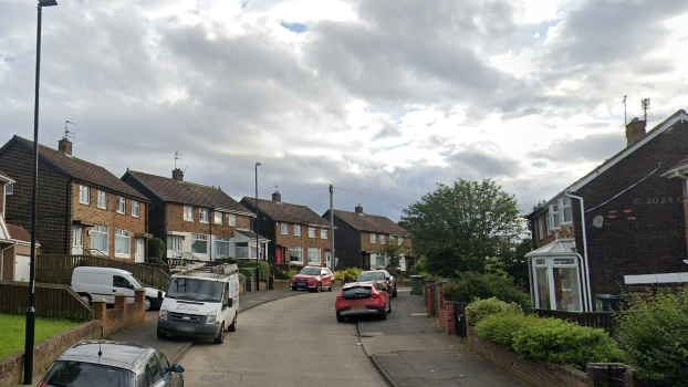 Rutherford Road, a curved street with filled with two-storey brick semi-detached houses. Several vehicles are parked half on the pavement along both sides of the street.