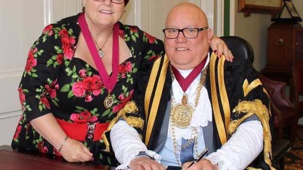 A man sitting down in mayoral garb and wearing glasses.