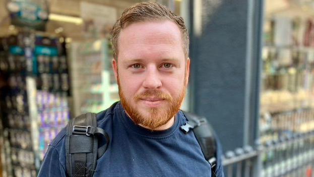 Luke is wearing a navy t-shirt, with a backpack and is looking into the camera. He has short ginger hair and a short beard, and stands in front of a shop window.