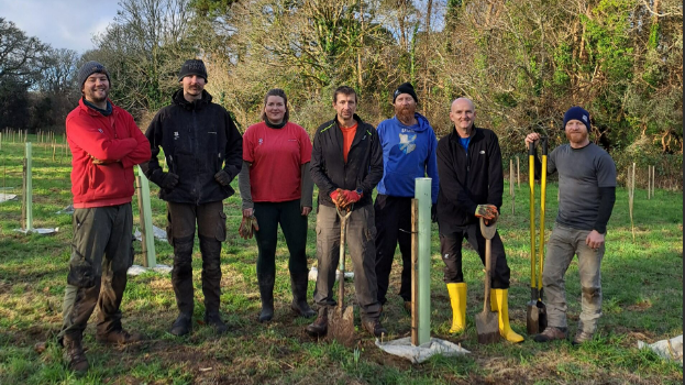 A group of people stood on a green field with trees behind them. Three of the people have spades and tools in their hands. All of the people in the photograph are smiling. 