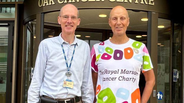 Professor Chris Parker on the left wearing blue shirt and hospital lanyard and Kevin Webber wearing a multi-coloured Royal Marsden Cancer Charity t-shirt standing outside entrance to Royal Marsden Hospital 