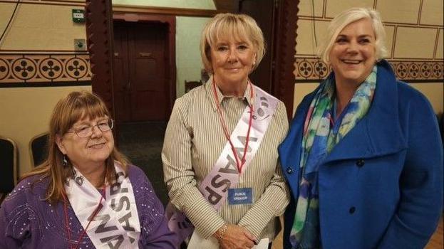 Three women, including Mary Smith (centre) looking at a camera. Two of the women are wearing WASPI branded sashes 