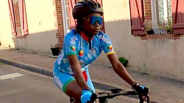 Georgette Vignonfodo, wearing a black cycle helmet, tinted sunglasses and light blue cycling lycras pedals down a street