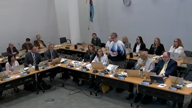 A still of Dacorum Borough Council's full meeting where many councillors are sitting around tables with laptops and microphones. A woman is standing up and reading from a piece of paper.