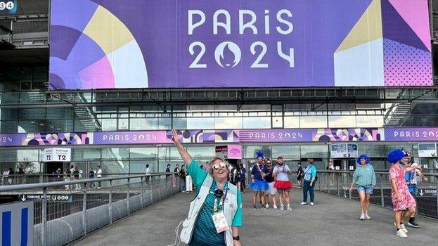 Karolyn Gaston at the sign for the Olympics - she is holding up her right hand doing  a peace sign 