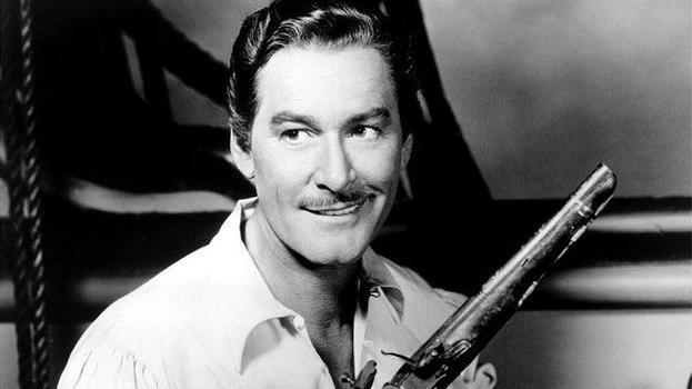 Errol Flynn with dark hair and a slight moustache wearing a light-coloured shirt and carrying a Blunderbuss-style gun in a black and white film still