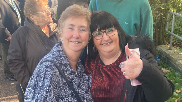 Pauline Tatham was stood on the left and was looking towards the camera with a light smile. She had short dark blonde hair and was wearing a black and white patterned coat. On the right was Biffy Roche who was looking towards the camera smiling with her thumb up. She had short black hair with a fringe and was wearing a red top and black coat.