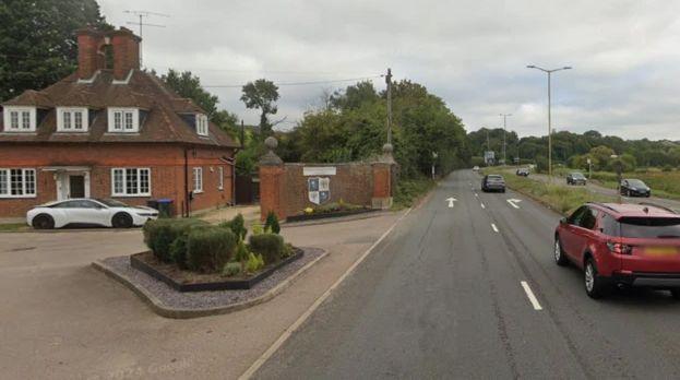 A dual carriageway which runs directly outside the entrance of a school drive which has some small hedges and a building at the end of it.