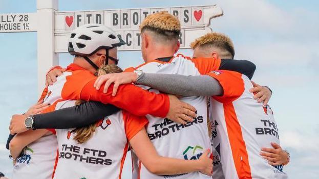 Four people hug with their backs to camera in front of the Land's End sign, which has been customised to say FTD Brothers. The man on the left of the picture is wearing a white bike helmet; a woman next to him has a blonde ponytail which can just be seen; the two men on the right have skin fade haircuts dyed blond on top. All people are wearing white and orange t-shirts saying The FTD Brothers.