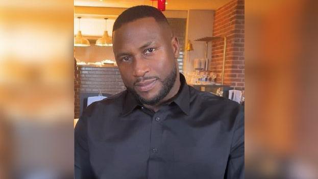 Theo Welsh wearing a black button-up shirt. He appears to be sitting in a restaurant with brick walls and a small fireplace behind him. He has short black hair which is shaven on the sides, brown eyes, and a dark trimmed beard. He is subtly smiling at the camera.