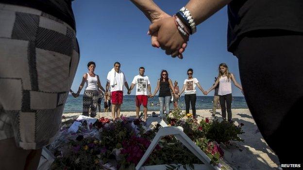 People hold hands as they pray in a circle around bouquets of flowers laid as mementos on the beach of the Imperial Marhaba resort