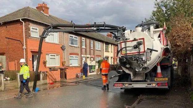 A water pumping tank truck pumps water from a street drain with two workers dressed in high visibility clothing and white hard hats stood next to it