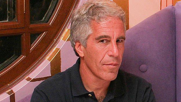 A close photo of Jeffrey Epstein. He sits down and looks at the camera, wearing a dark coloured shirt